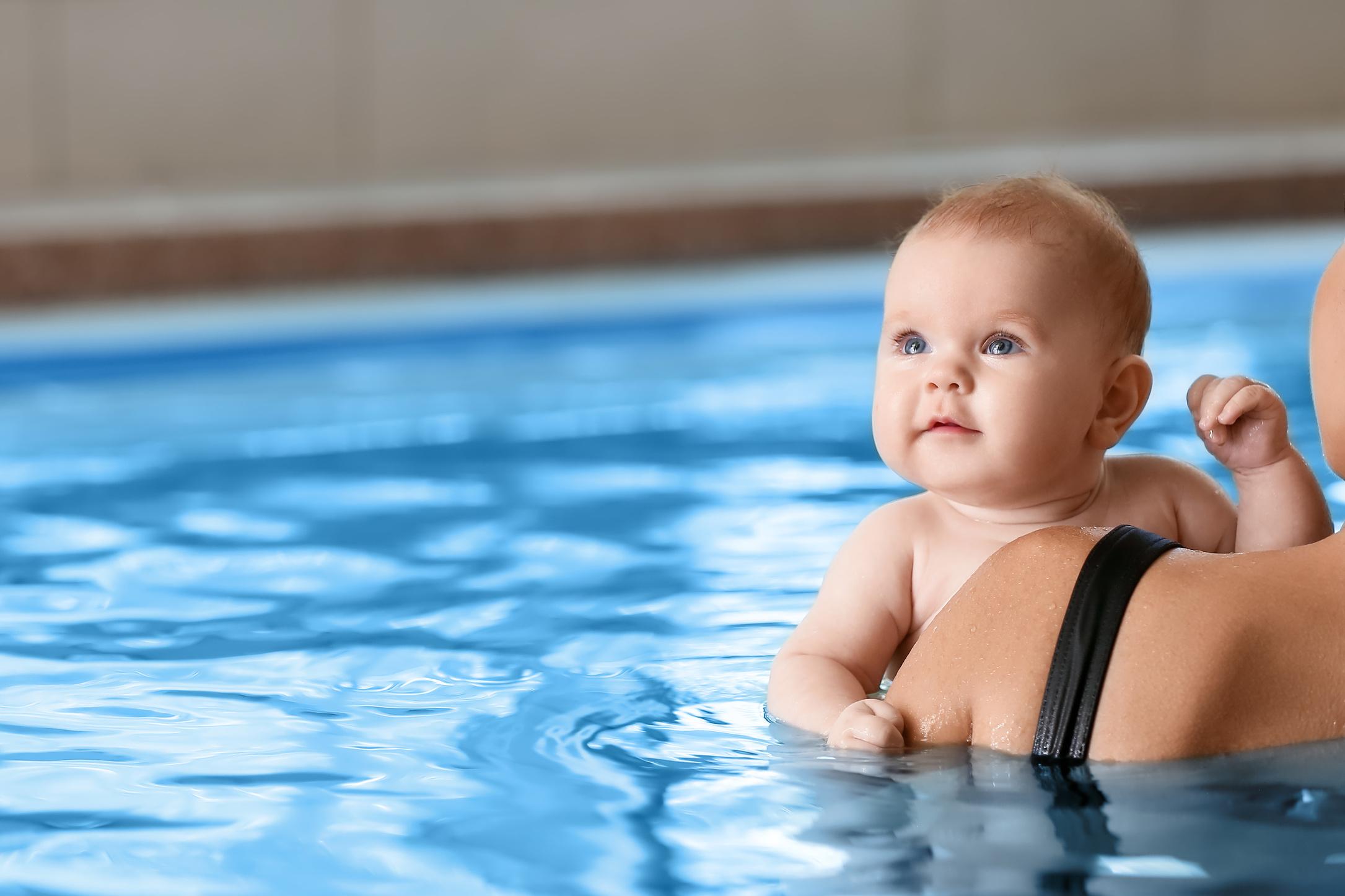 Baby mit blauen Augen, das in einem Schwimmbad auf dem Arm eines Erwachsenen schwimmt. Wasser spiegelt sich sanft.