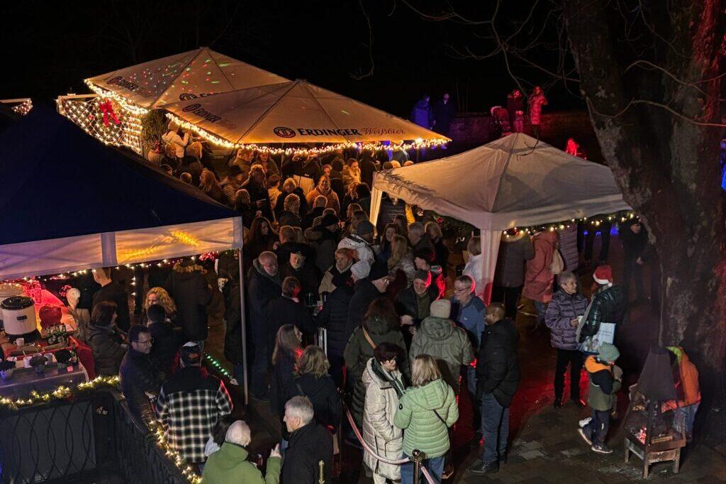 Menschenmenge auf einem festlichen Weihnachtsmarkt nachts, umgeben von beleuchteten Ständen und Dekorationen.