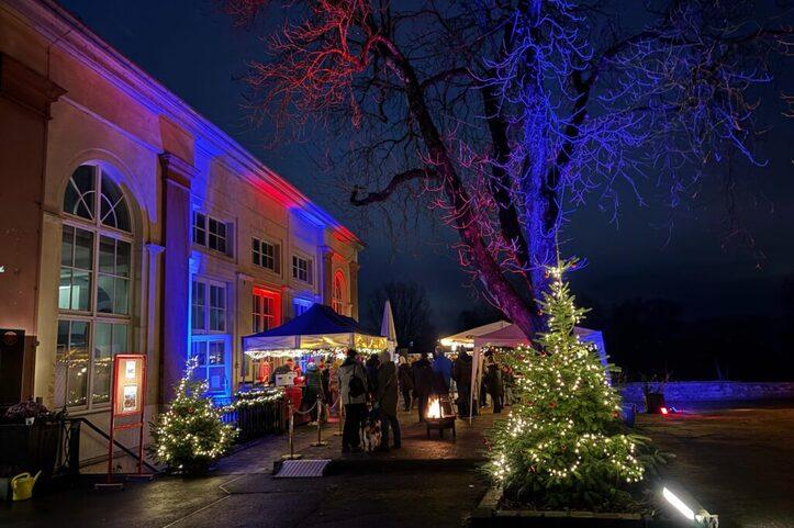 Beleuchteter Au&szlig;enbereich eines Weihnachtsmarkts mit St&auml;nden, einem geschm&uuml;ckten Baum und Besuchern am Abend.