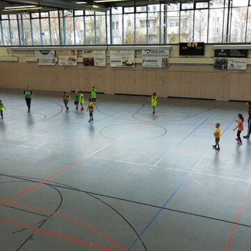 Kinder spielen in einer Sporthalle Fu&szlig;ball, umgeben von Fenstern und W&auml;nden mit Werbebannern.