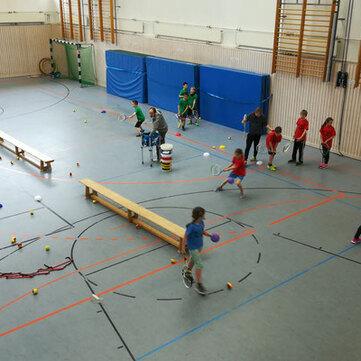Gruppen von Kindern in Sportkleidung spielen in einer Turnhalle mit verschiedenen B&auml;llen und Sportger&auml;ten.