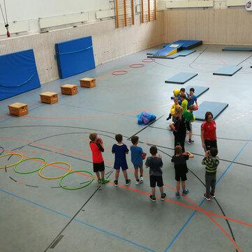 Kindersportgruppe mit mehreren Kindern in einer Turnhalle, diverse Sportger&auml;te und &Uuml;bungsstationen sichtbar.