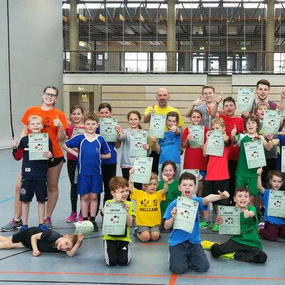 Gruppenfoto von 30 Kindern in Sportkleidung, die stolz ihre Urkunden in einer Turnhalle pr&auml;sentieren.
