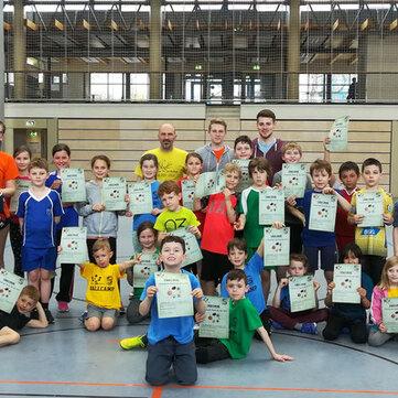 Gruppenfoto von Kindern und Betreuern in sportlichen Shirts, alle halten Urkunden in einer Sporthalle.