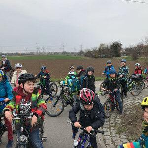 Gruppe von Kindern in Radhelmen auf Fahrr&auml;dern, versammelt auf einem Feldweg mit grauem Himmel im Hintergrund.