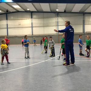 Trainer erkl&auml;rt Fu&szlig;balltechnik einer Gruppe von Kindern in Sportkleidung in einer Turnhalle.