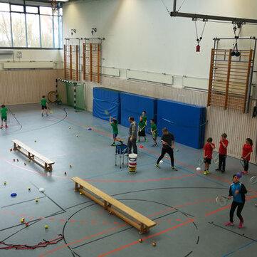 Gruppe von Kindern in einer Sporthalle beim Tennis-Training, umgeben von B&auml;llen und &Uuml;bungsmaterialien.