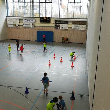 Kinder spielen in einer Sporthalle, umringt von H&uuml;tchen und einem Tor; einige Erwachsene beaufsichtigen das Training.