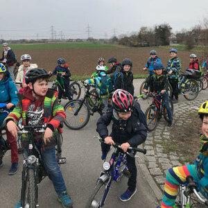 Gruppe von Kindern in Radhelmen auf Fahrr&auml;dern, versammelt auf einem Feldweg mit grauem Himmel im Hintergrund.
