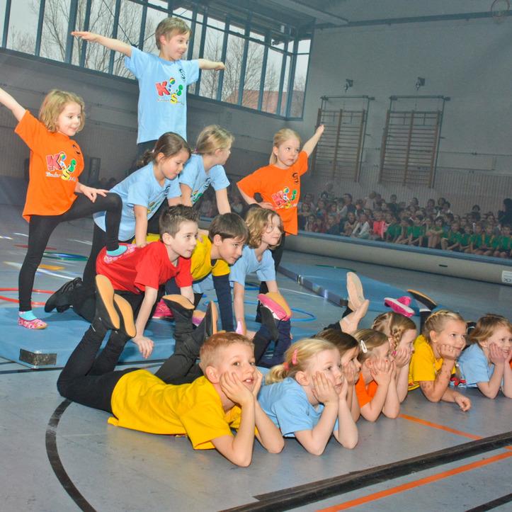 Gruppen von Kindern in bunten T-Shirts stehen und liegen auf Matten, w&auml;hrend sie einander unterst&uuml;tzend anfeuern.