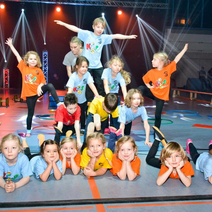 Kinder in farbigen T-Shirts machen eine kreative Gruppenchoreografie auf einer Sportfl&auml;che mit B&uuml;hnenbeleuchtung.