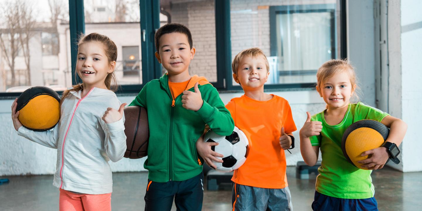Vier Kinder stehen in einem Sportraum und halten verschiedene B&auml;lle, w&auml;hrend sie l&auml;cheln und Daumen hoch zeigen.