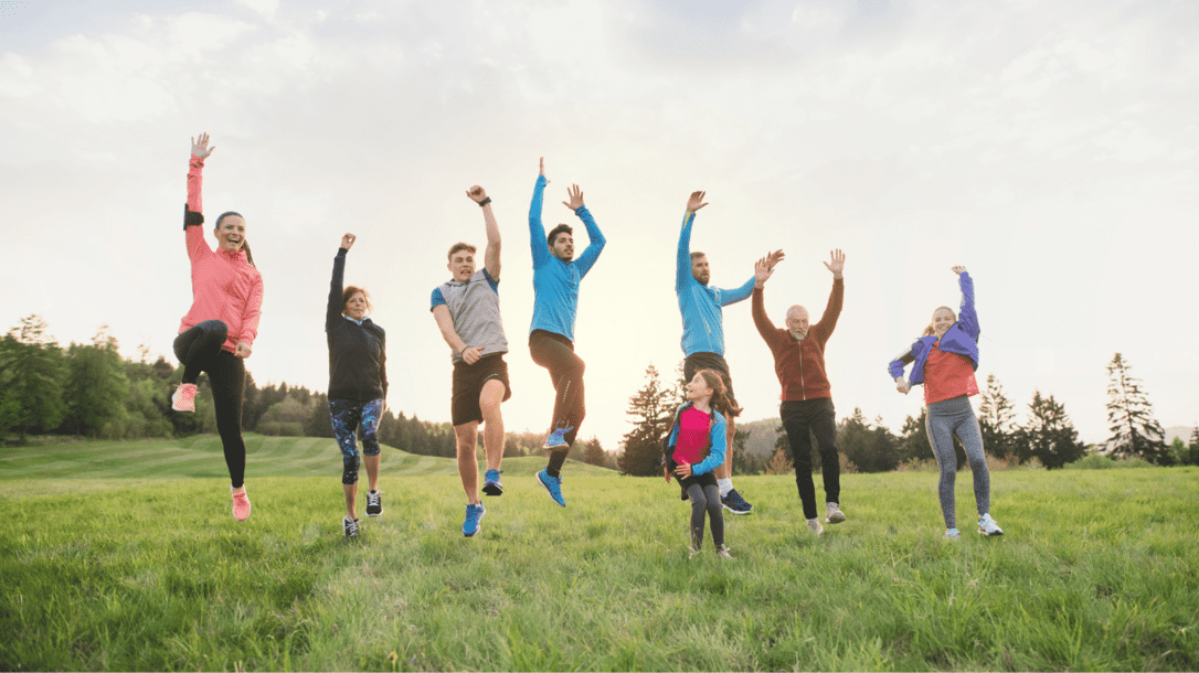 Gruppe von acht Personen springt fr&ouml;hlich auf einer Wiese vor einem bewaldeten Hintergrund in der Abendd&auml;mmerung.