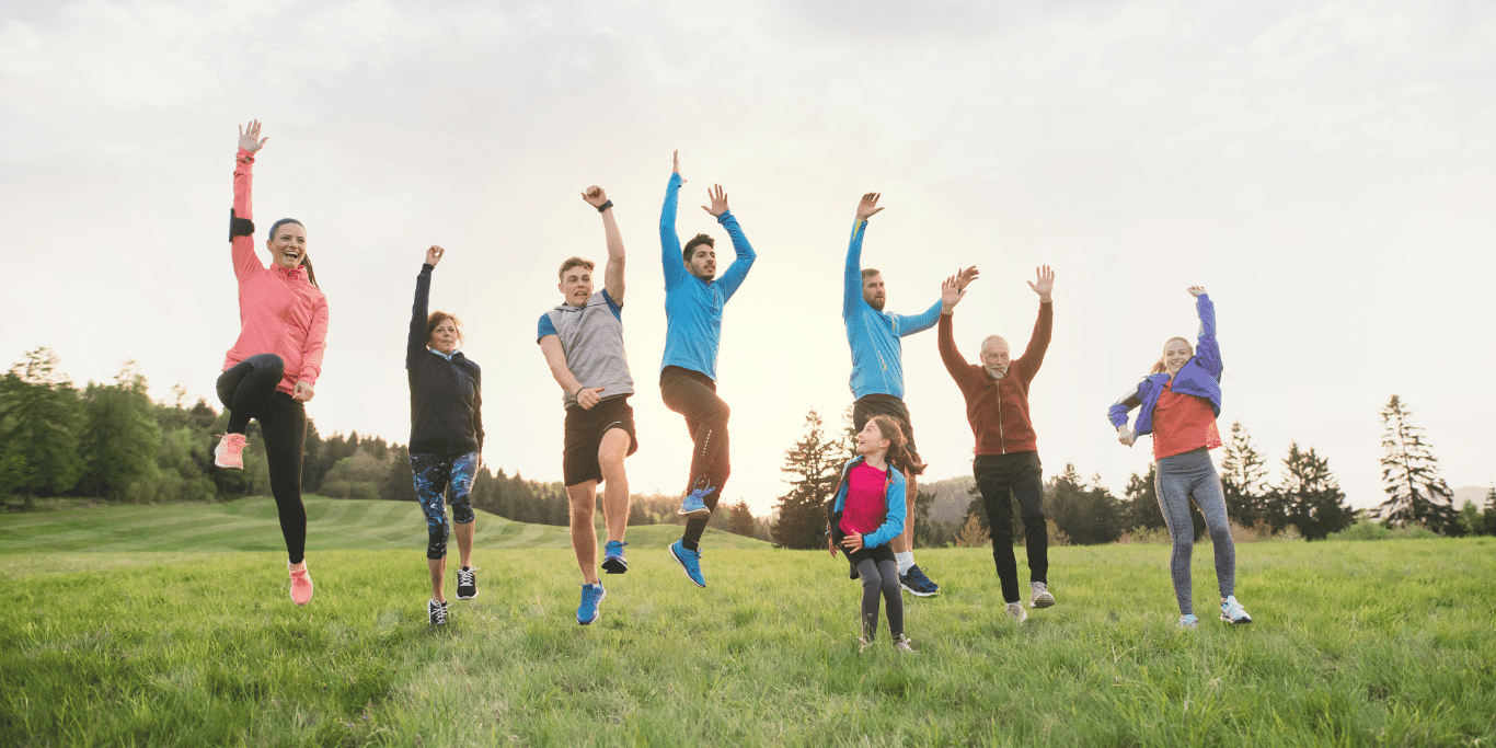 Gruppe von acht Personen springt fr&ouml;hlich auf einer Wiese vor einem bewaldeten Hintergrund in der Abendd&auml;mmerung.
