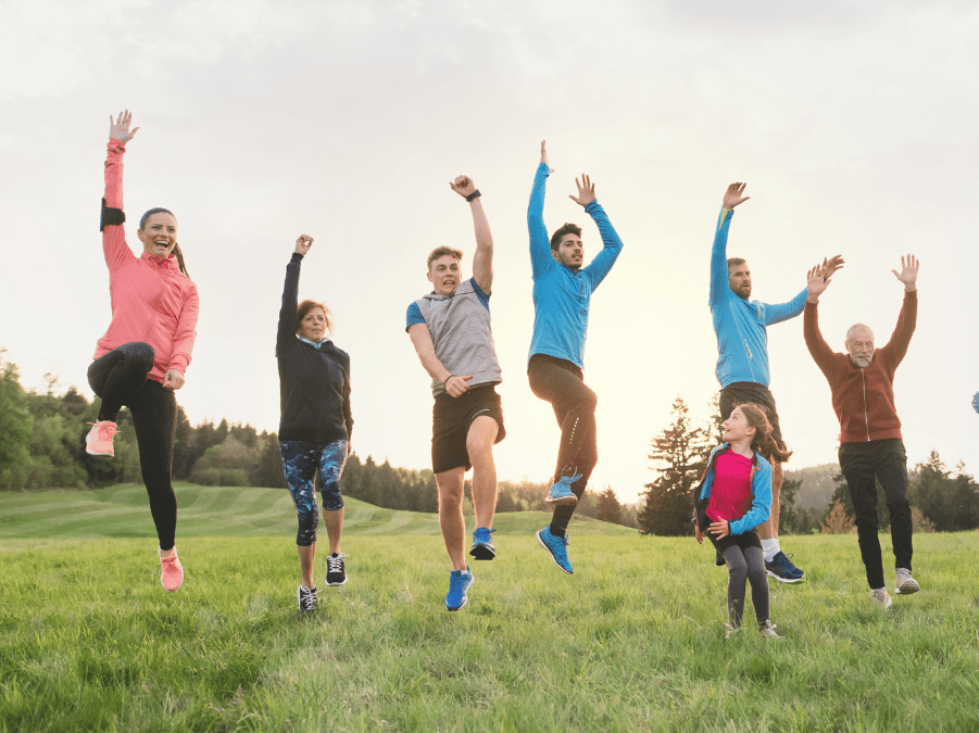Gruppe von acht Personen springt fr&ouml;hlich auf einer Wiese vor einem bewaldeten Hintergrund in der Abendd&auml;mmerung.