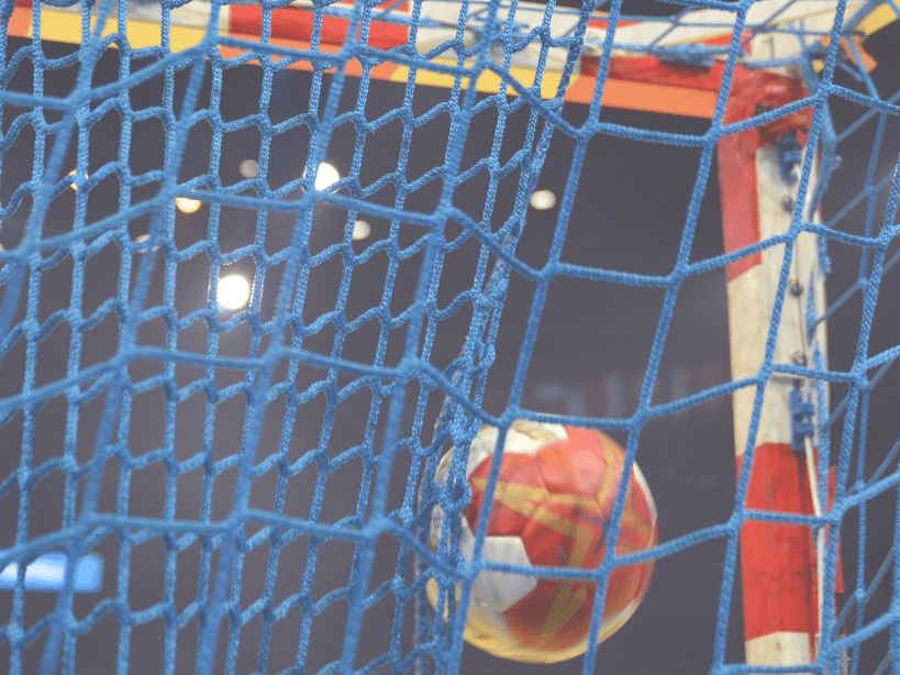 Nahaufnahme eines Handballs, der ins Netz eines Torrahmens aus blauen und wei&szlig;en Schn&uuml;ren fliegt.