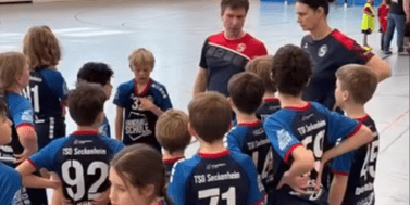 Gruppe von Kindern in Sportkleidung um einen Trainer in der Halle, w&auml;hrend sie Anweisungen erhalten.