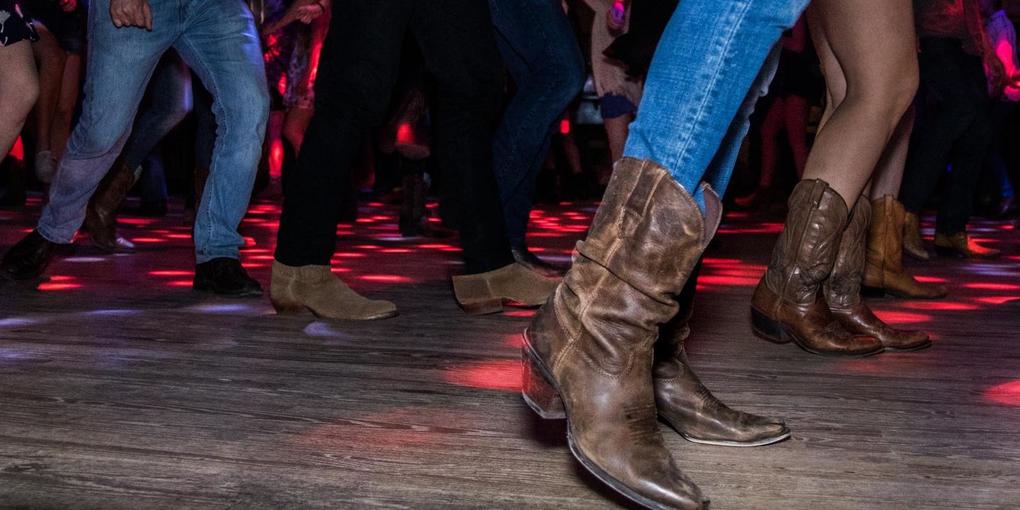 Tanzende F&uuml;&szlig;e in Cowboy-Stiefeln auf einer Holzbodenfl&auml;che, umgeben von bunten Lichtpunkten.