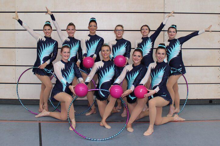 Gruppe von zehn Turnerinnen in glitzernden Kost&uuml;men, die bunte B&auml;lle und Reifen halten, posieren auf einer Sportfl&auml;che.