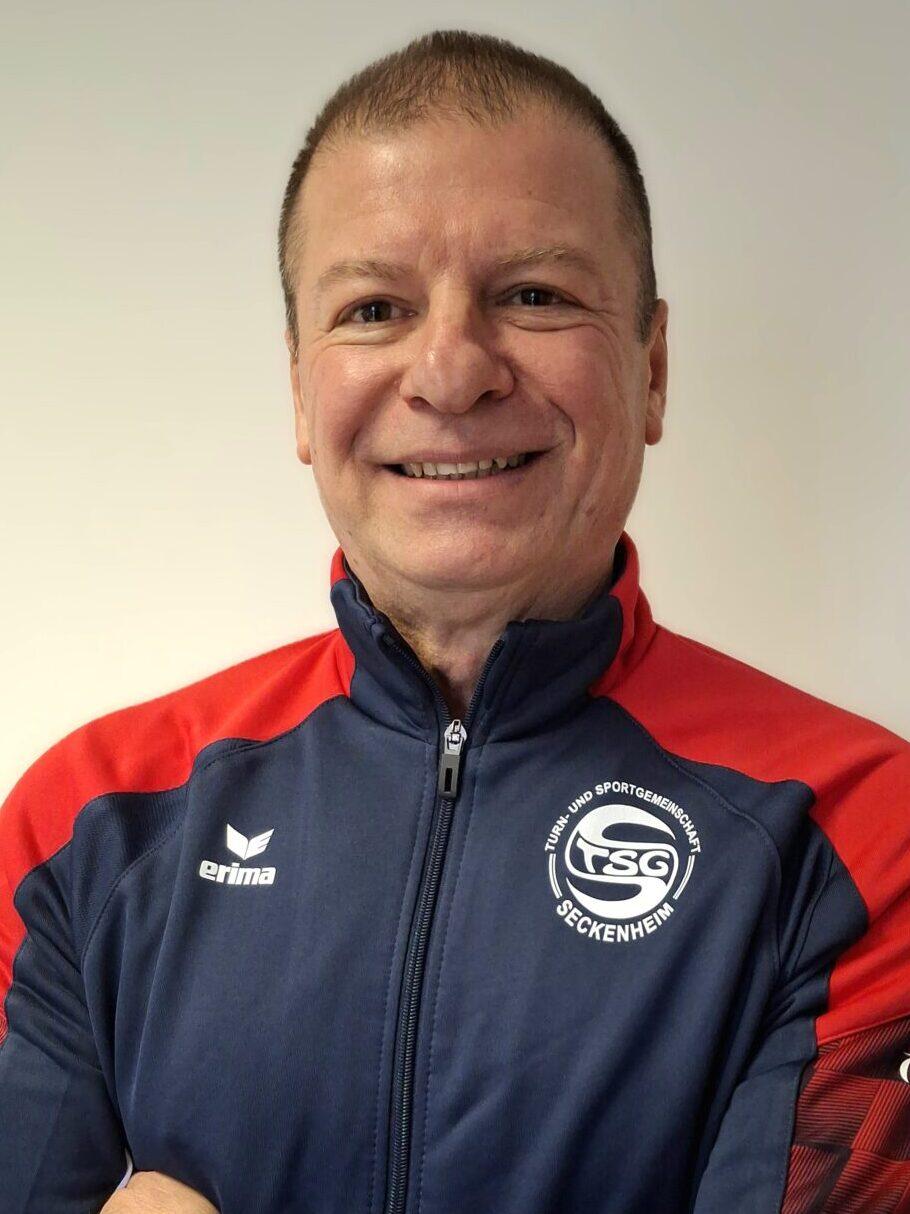 L&auml;chelnder Mann mit verschr&auml;nkten Armen, tr&auml;gt einen blauen Trainingsanzug mit roten Akzenten und Logo.
