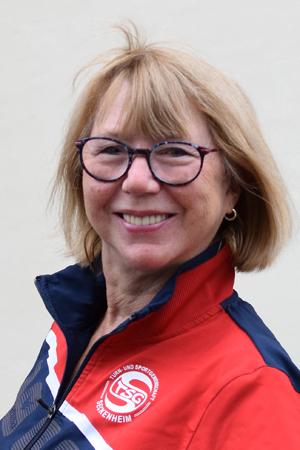 Frau mit blonden, schulterlangen Haaren und Brille tr&auml;gt eine rot-blau gestreifte Sportjacke und l&auml;chelt freundlich.