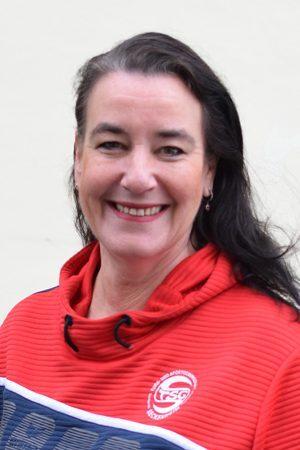 L&auml;chelnde Frau mit langen, dunklen Haaren in rotem Sportoberteil, vor einem hellen Hintergrund.