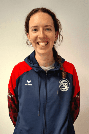 L&auml;chelnde Frau mit langen Haaren in einem Sportanzug vor neutralem Hintergrund.