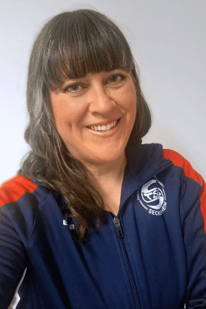 Portr&auml;t einer l&auml;chelnden Frau mit langen, glatten Haaren, die einen dunkelblauen Sportanzug tr&auml;gt.