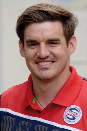 L&auml;chelnder Mann mit kurzen Haaren tr&auml;gt ein rotes Poloshirt und steht vor einer hellen Hintergrundwand.