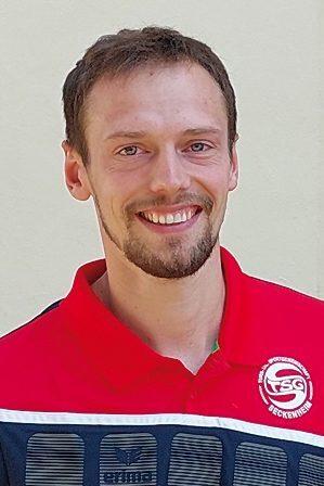L&auml;chelnder Mann in rotem Sporttrikot mit Logo der TSG Beckenheim vor einfarbiger Wand.