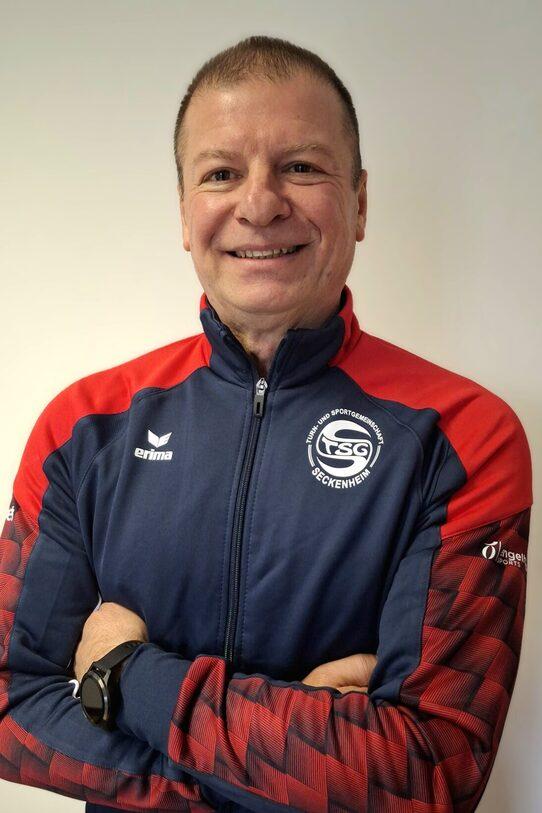 Lächelnder Mann mit verschränkten Armen, trägt einen blauen Trainingsanzug mit roten Akzenten und Logo.