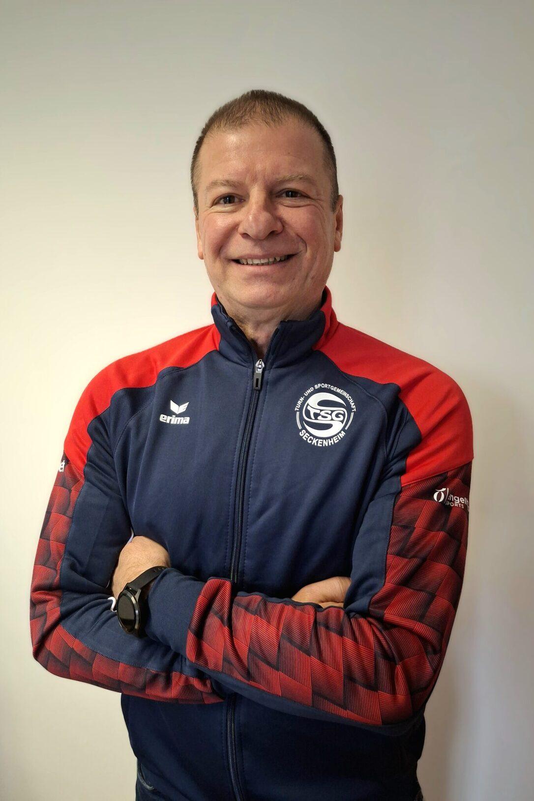 L&auml;chelnder Mann mit verschr&auml;nkten Armen, tr&auml;gt einen blauen Trainingsanzug mit roten Akzenten und Logo.