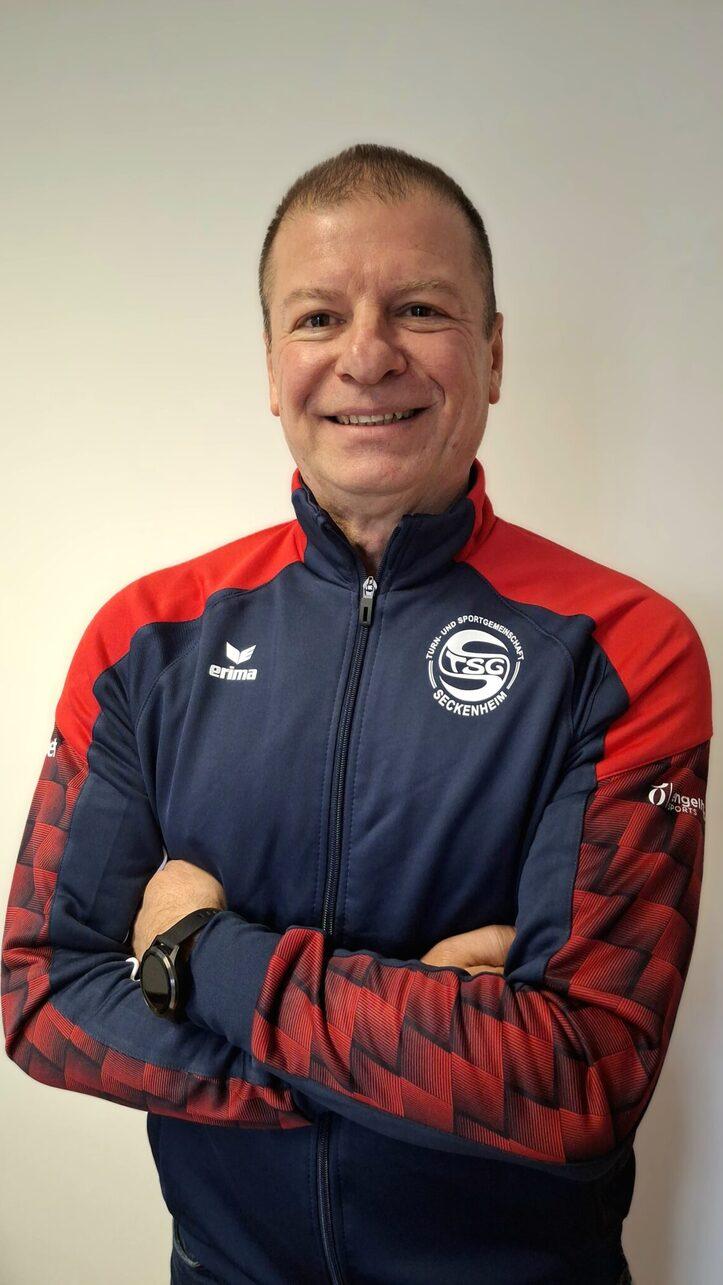 L&auml;chelnder Mann mit verschr&auml;nkten Armen, tr&auml;gt einen blauen Trainingsanzug mit roten Akzenten und Logo.