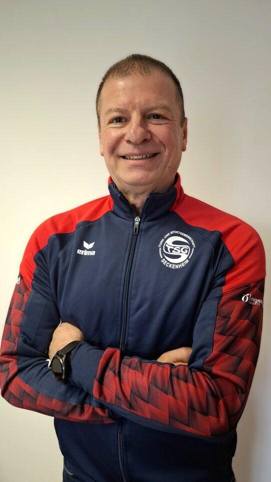 L&auml;chelnder Mann mit verschr&auml;nkten Armen, tr&auml;gt einen blauen Trainingsanzug mit roten Akzenten und Logo.