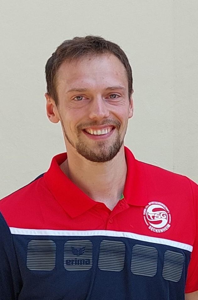 L&auml;chelnder Mann mit Kurzhaarschnitt tr&auml;gt ein rotes Sportshirt mit TSG-Logo vor einer einfarbigen Wand.