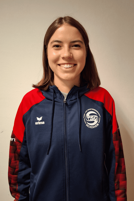 Portr&auml;t einer jungen Frau in einer blauen Sportjacke mit rotem &Auml;rmel und Logo der TSG Seckenheim. Sie l&auml;chelt.
