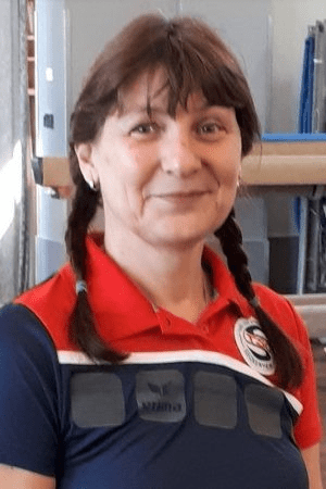 Frau mit Zopf trägt ein rotes Sportshirt mit Emblem und lächelt in einem Innenraum mit Sportequipment.