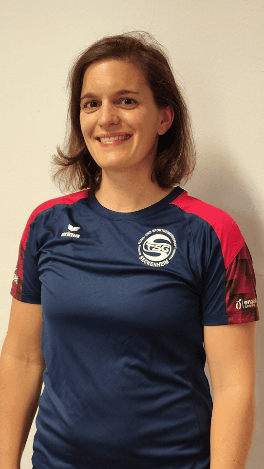 Frau mit kurz geschnittenem Haar tr&auml;gt ein blaues Sportshirt mit rot-schwarzen &Auml;rmeln und l&auml;chelt vor einer wei&szlig;en Wand.