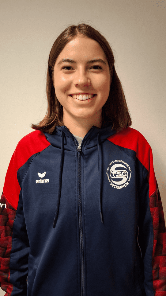 Portr&auml;t einer jungen Frau in einer blauen Sportjacke mit rotem &Auml;rmel und Logo der TSG Seckenheim. Sie l&auml;chelt.