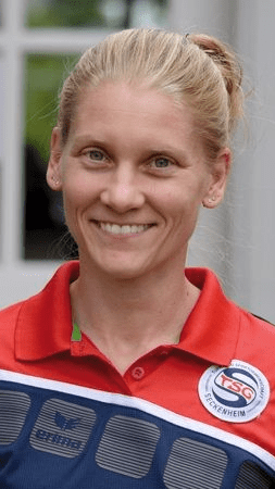 L&auml;chelnde Frau in rotem Sporttrikot mit Vereinslogo, steht vor einer Fensterfront.