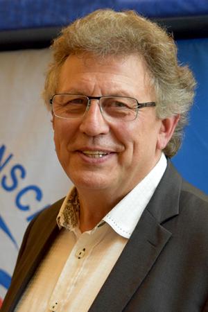 Portr&auml;t eines l&auml;chelnden Mannes mit Brille, grauen Haaren und einem Anzug, vor einem blauen Hintergrund.