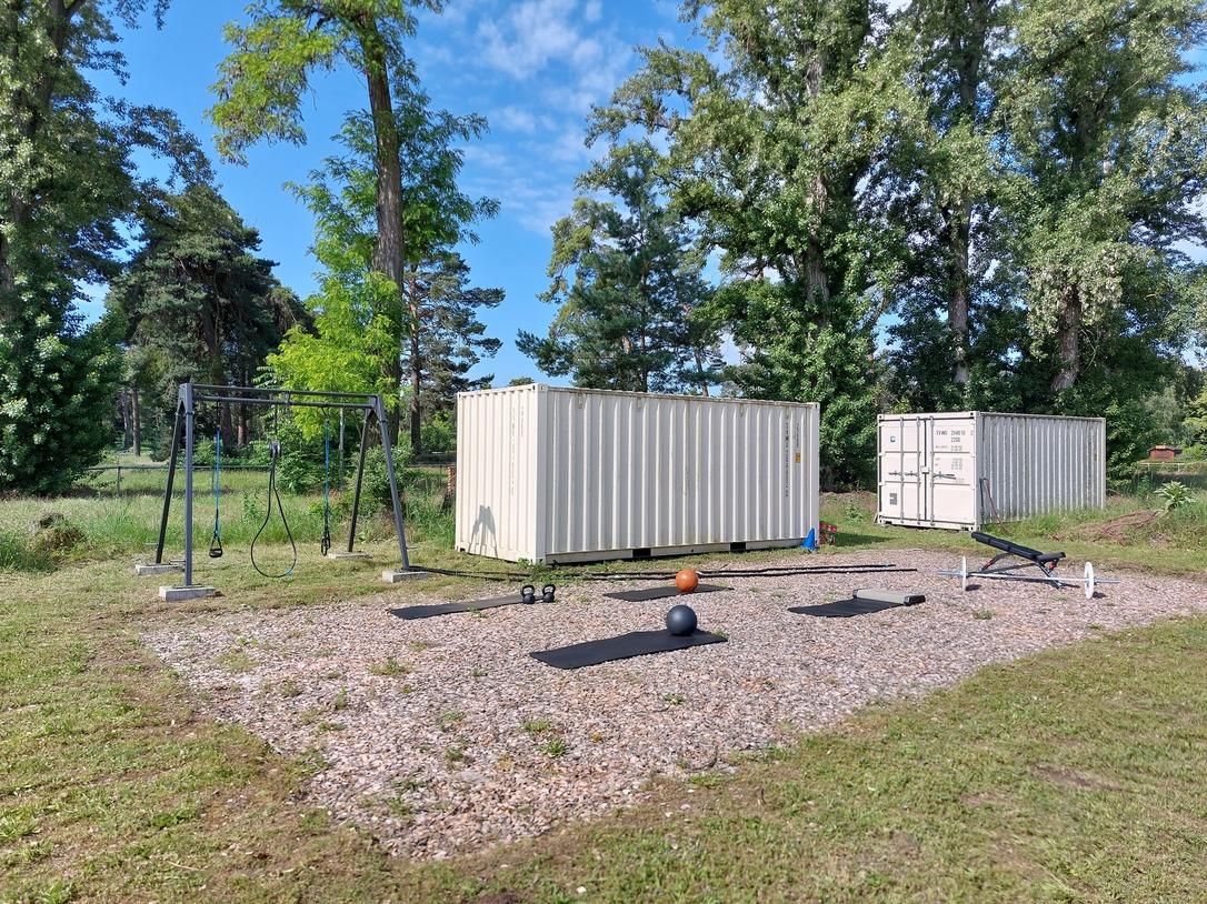 Fitnessbereich im Freien mit Trainingsger&auml;ten, Matten und zwei Containern auf einer Wiese umgeben von B&auml;umen.