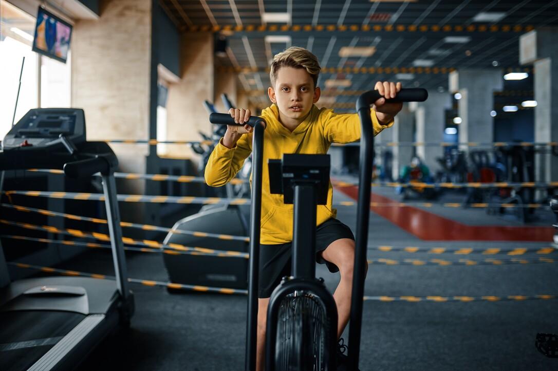 Ein Junge in einem gelben Hoodie trainiert auf einem station&auml;ren Fahrrad in einem Fitnessstudio.