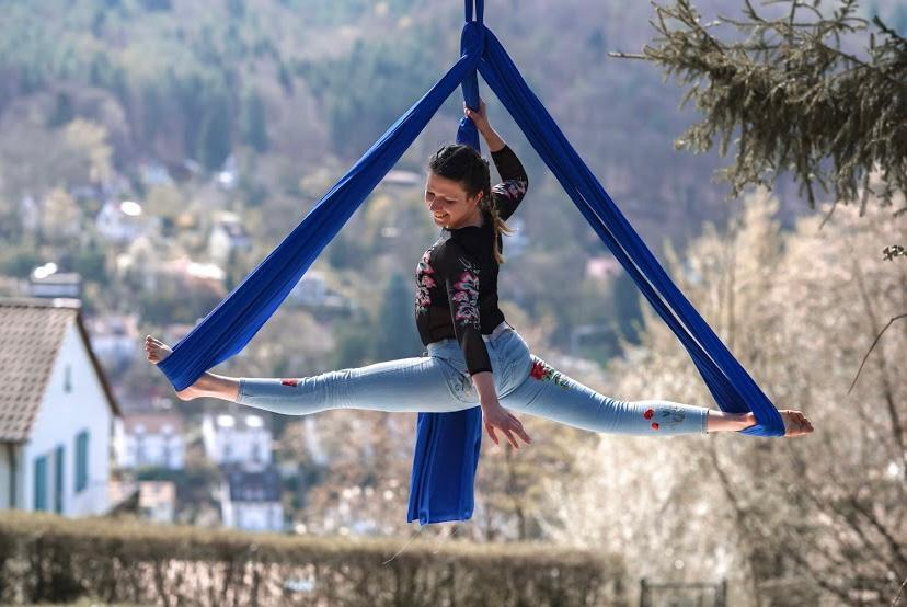Frau in schwarzen Oberteil und Jeans &uuml;bt Aerial Silk in blauem Tuch vor einer nat&uuml;rlichen Landschaft.