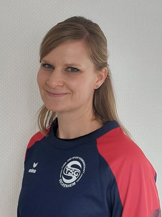Portr&auml;t einer l&auml;chelnden Frau mit blonden Haaren, in einem Sportshirt mit einem Logo im Hintergrund.