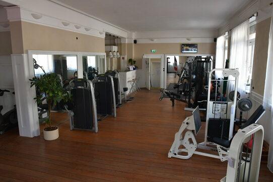 Fitnessstudio mit mehreren Trainingsgeräten und großen Spiegeln, Holzfußboden und Tageslicht durch Fenster.