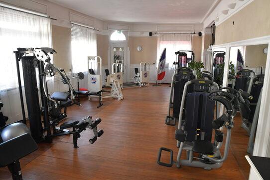 Fitnessraum mit verschiedenen Kraftger&auml;ten, h&ouml;lzernem Boden und gro&szlig;en Fenstern, die Tageslicht hereinlassen.