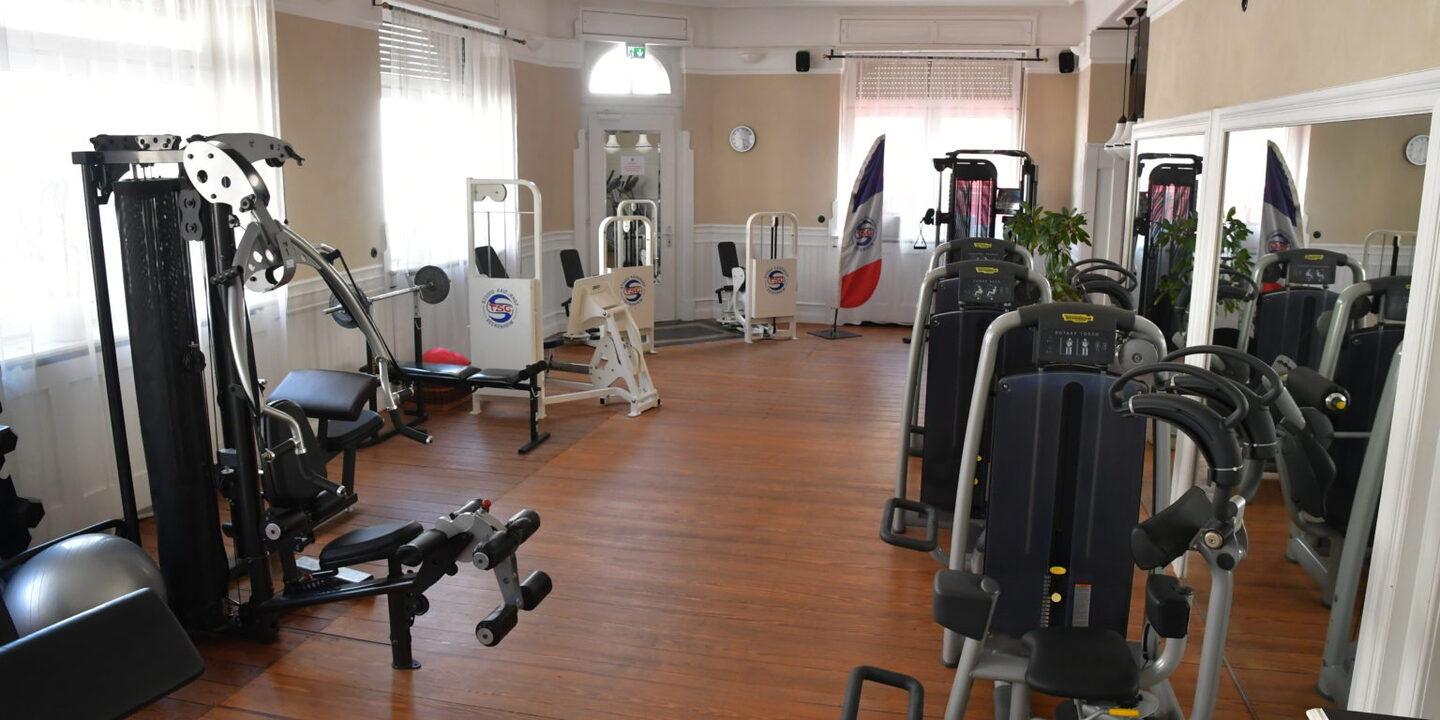 Fitnessraum mit verschiedenen Kraftger&auml;ten, h&ouml;lzernem Boden und gro&szlig;en Fenstern, die Tageslicht hereinlassen.