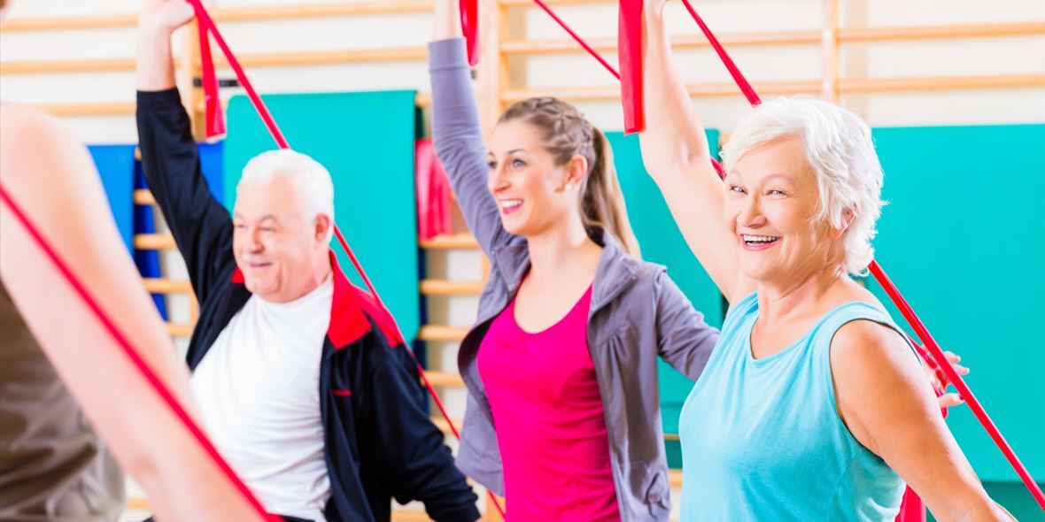 Gruppe von Menschen im Fitnessraum, die mit roten Widerstandsb&auml;ndern &Uuml;bungen durchf&uuml;hren und l&auml;cheln.