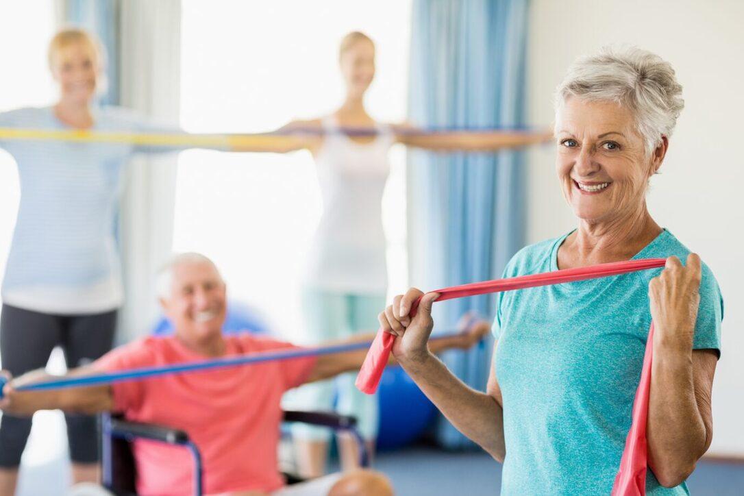 &Auml;ltere Frau h&auml;lt ein rotes Widerstandsband, w&auml;hrend andere im Hintergrund mit elastischen B&auml;ndern trainieren.